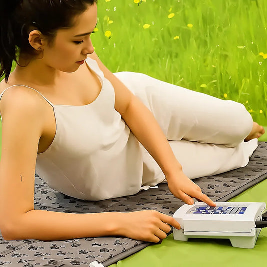 QRS 101 PEMF therapy device with control panel, used on a gray mat in a grassy setting by a woman in a light outfit.