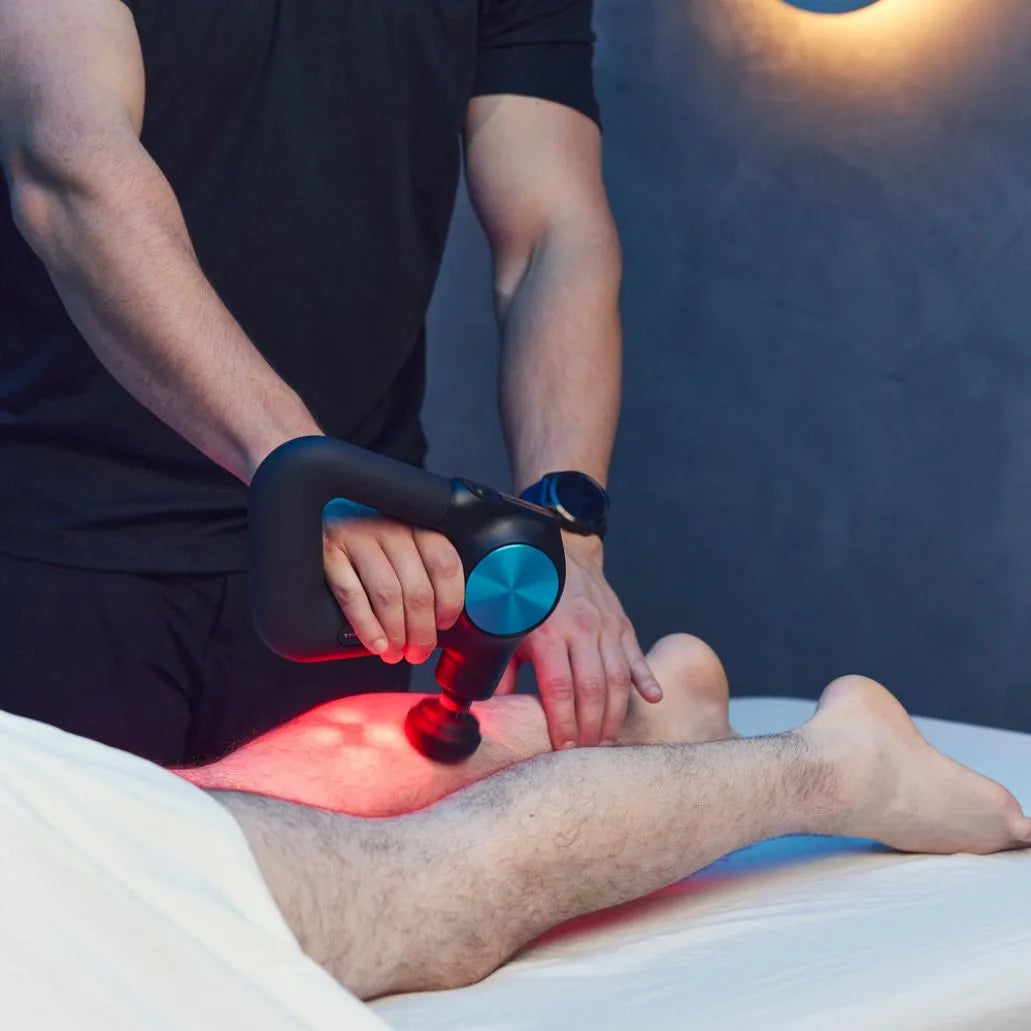 Person using a massage gun on another person's leg in a clinical setting.